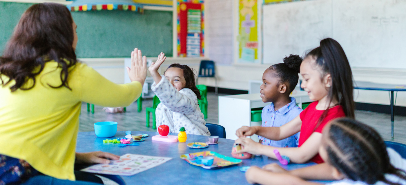 teacher giving a high five to a female child with other female children looking on in a classroom