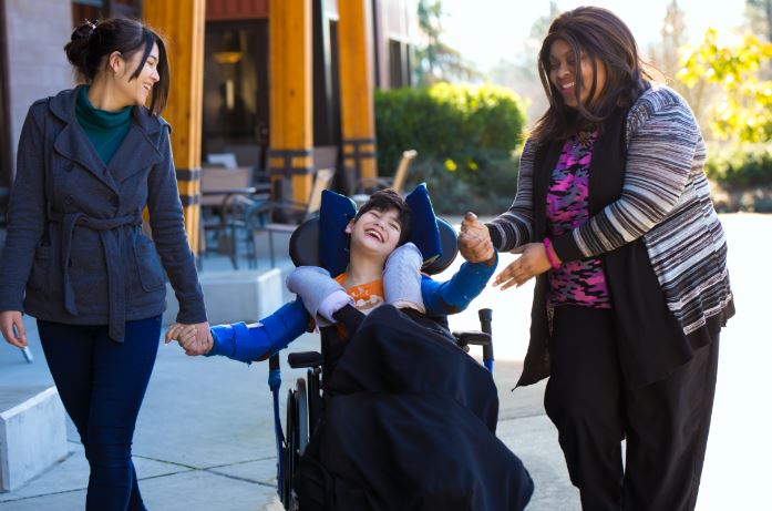 special needs child in a wheelchair in the middle with a woman on each side holding the child\'s hand