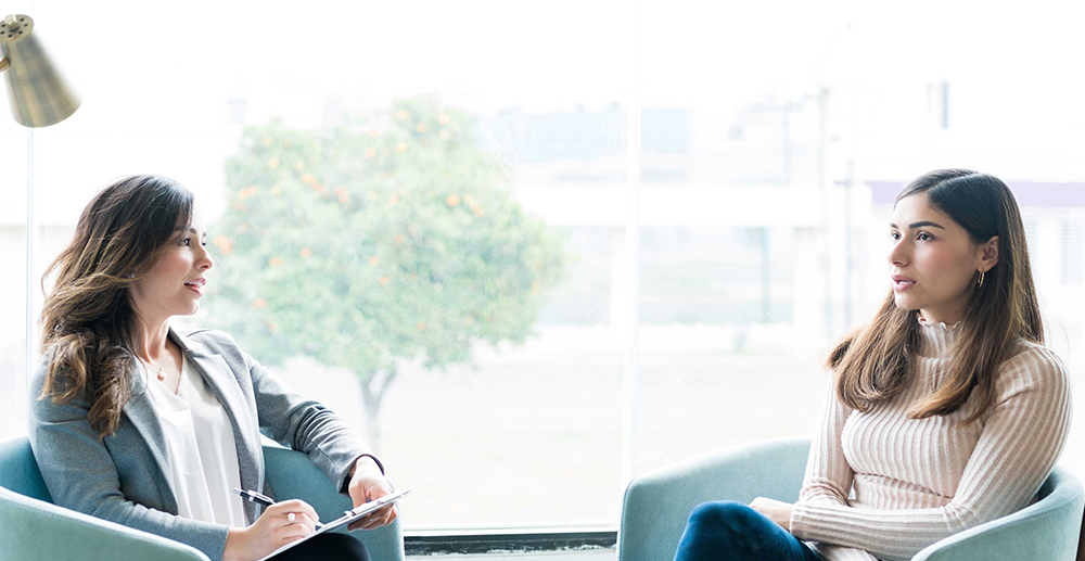 two women with long hair sitting on chairs angled towards each other. The woman on right is talking and woman on left has a notepad and pen on her lap. Window to outside in the background. 