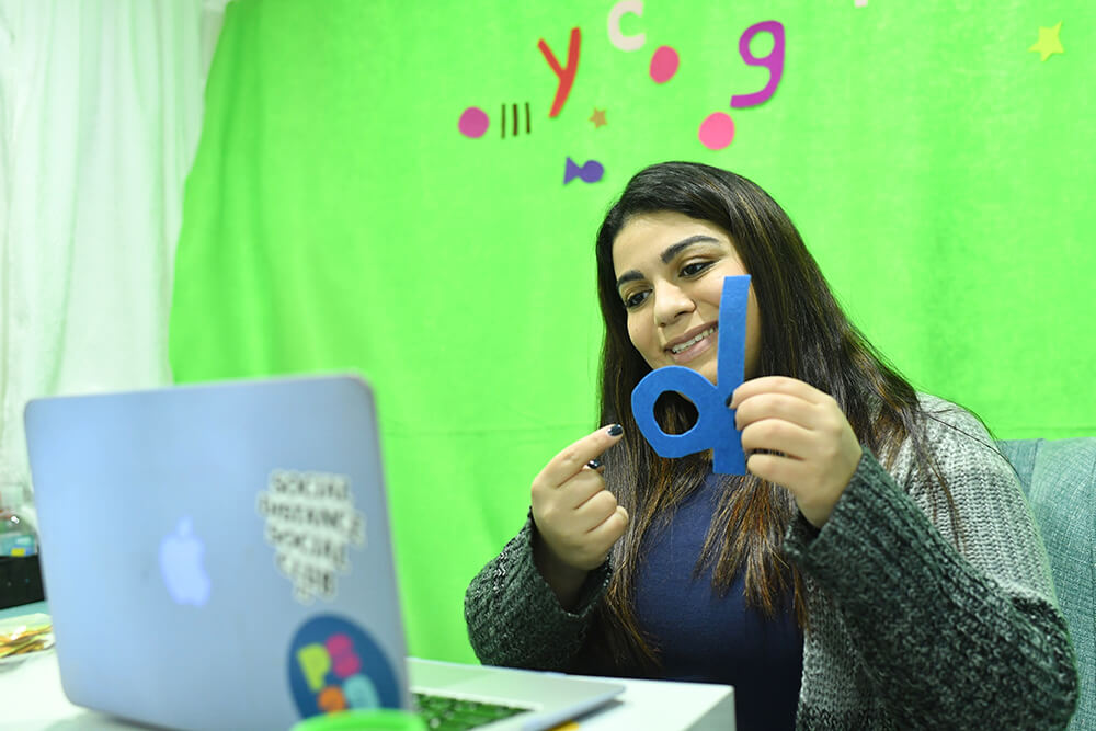 Tammy Youseflaleh, sitting in front of a laptop, talking to her students through zoom, holding and pointing to a "d" with a green screen with letters and shapes behind her.