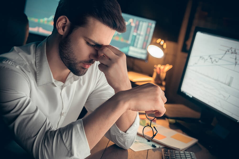 A man looking worried or depressed sitting at his desk in front of a computer with another computer screen in background