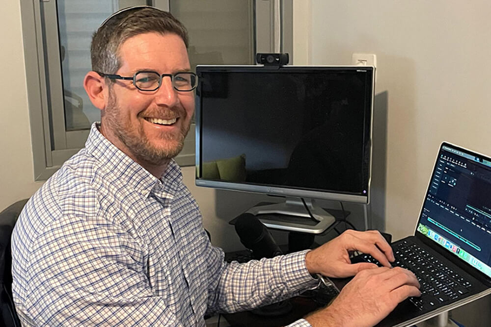 Ari Ginsberg, an alum of Touro University\'s School of Health Sciences, types on a laptop while sitting beside another computer.